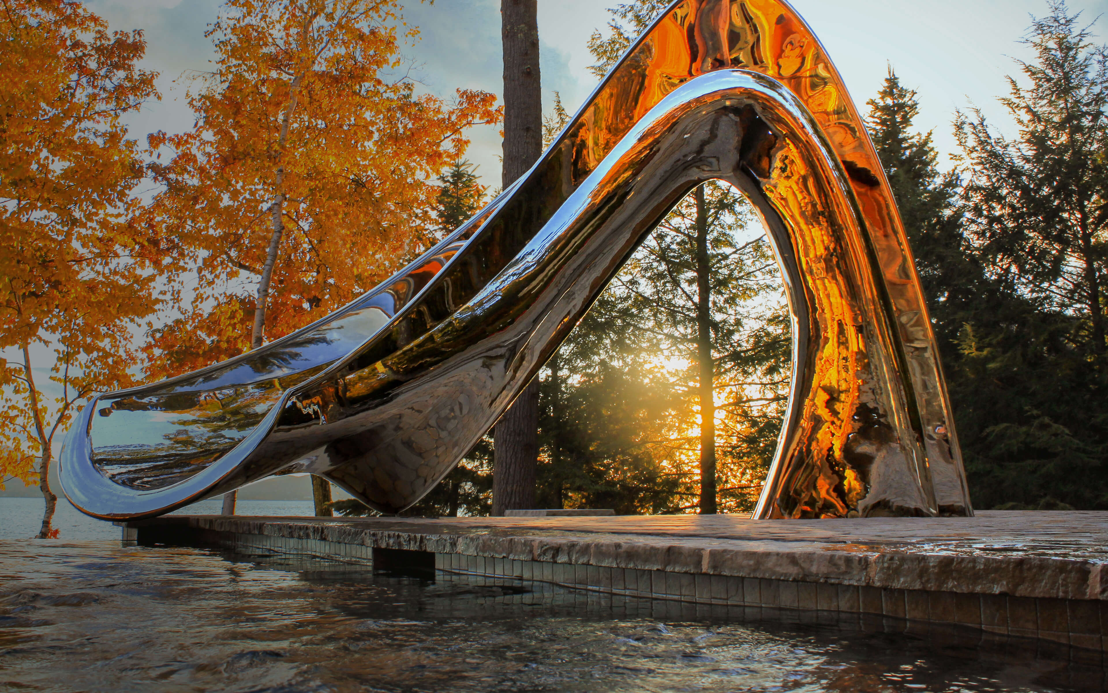 Sculptural pool slide in mirror polished metal next to lake at sunset, in collaboration with water shape designer