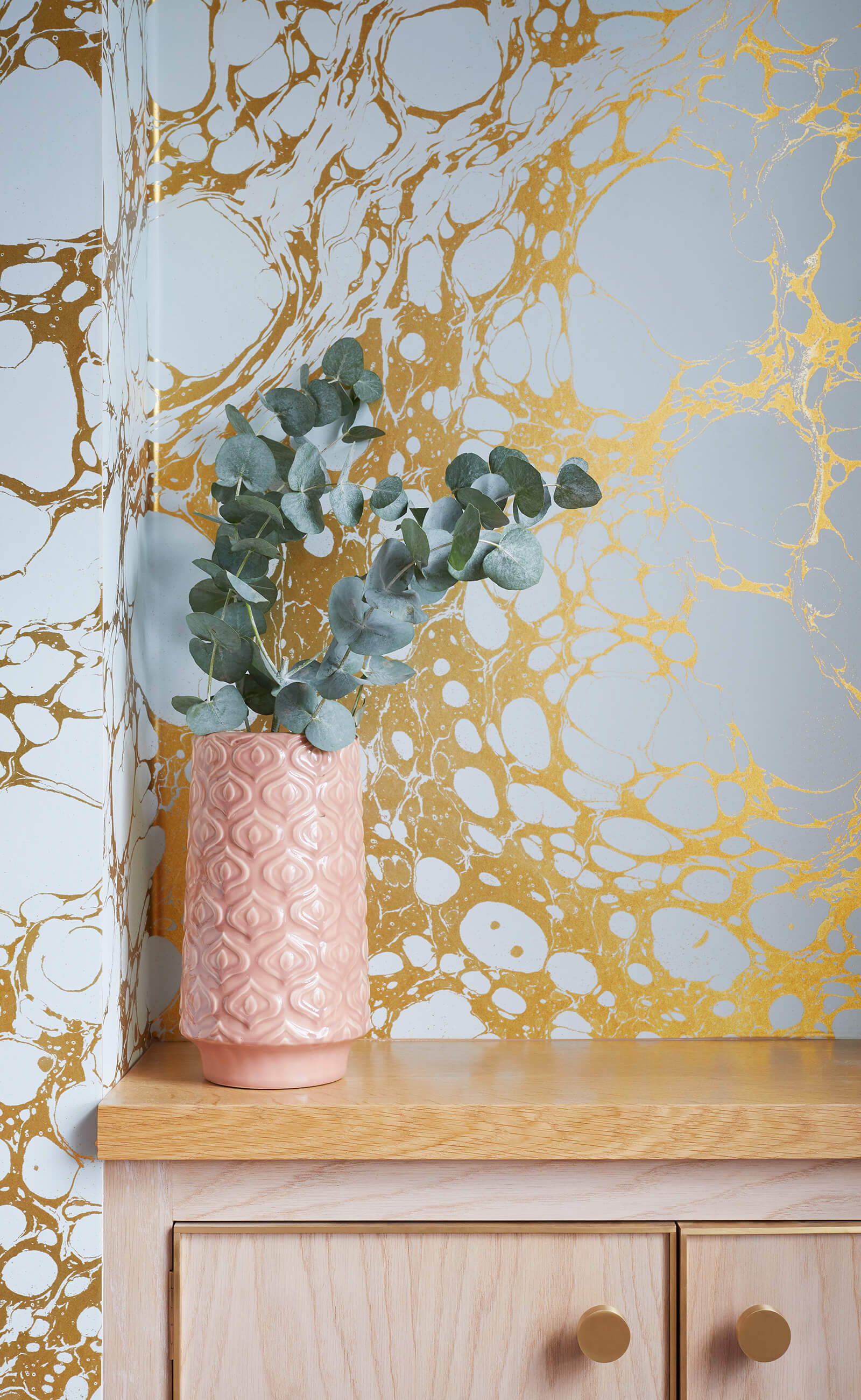 Detail of soft pink bedside cabinet with brass details next to gold and grey marbled wallpaper