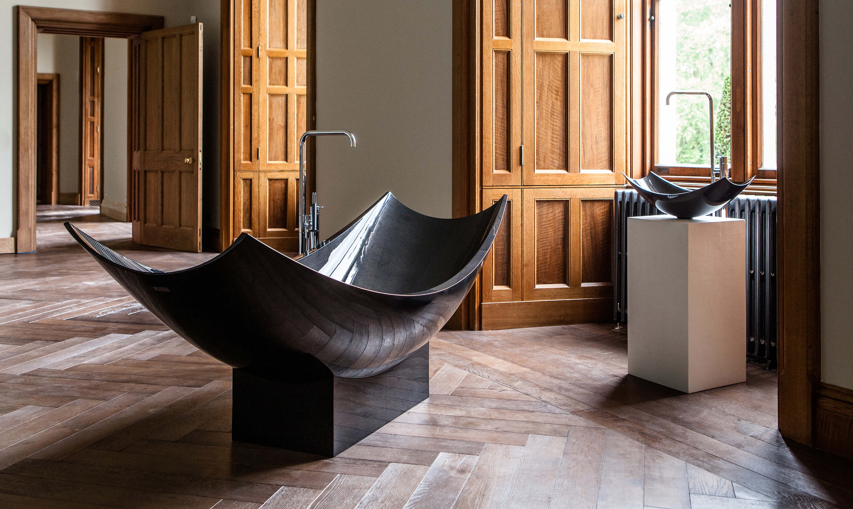 A black freestanding bateau bath tub in carbon fibre in a panelled room with black basin.