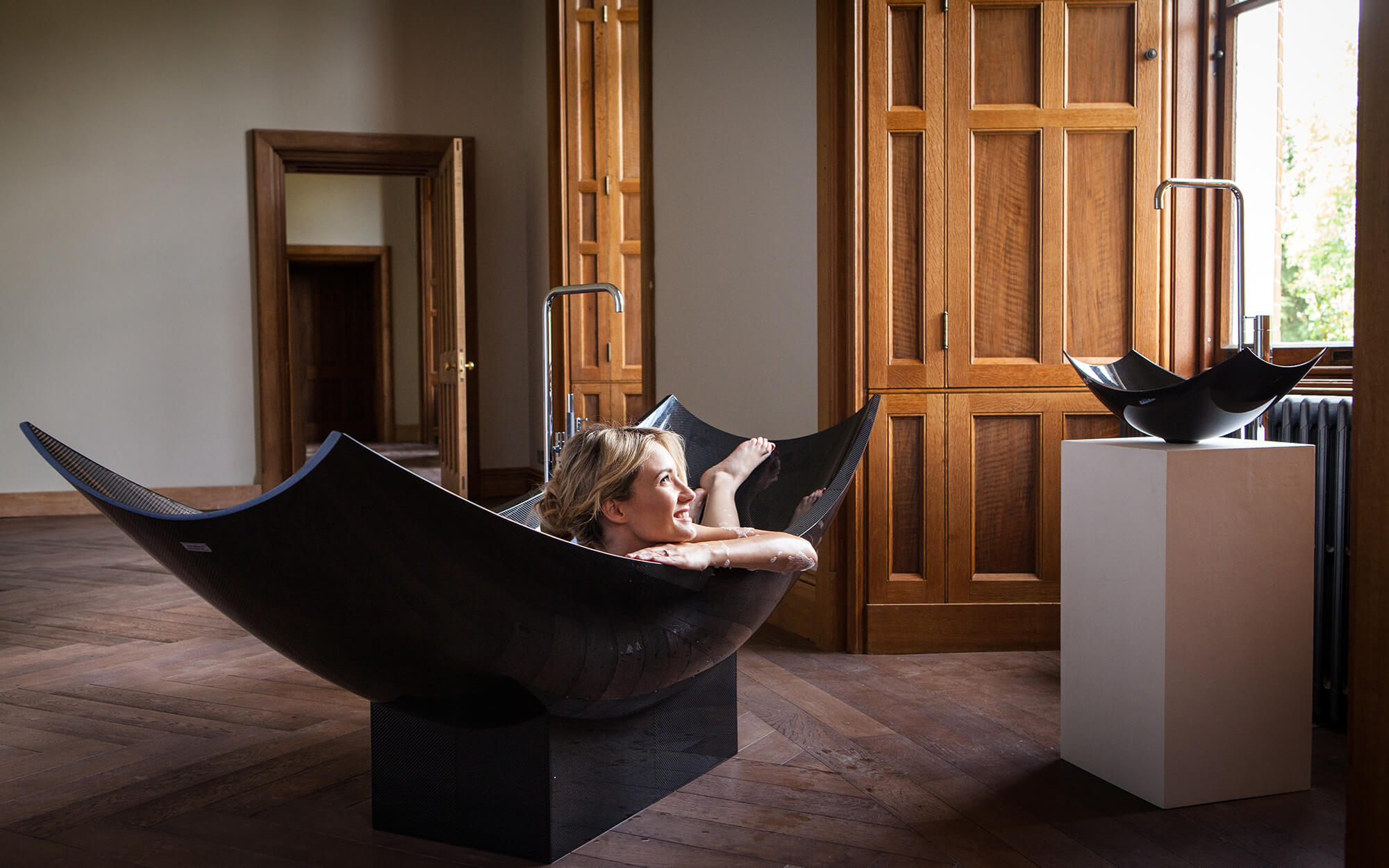 Woman lying in black freestanding bath on wooden floor next to window.