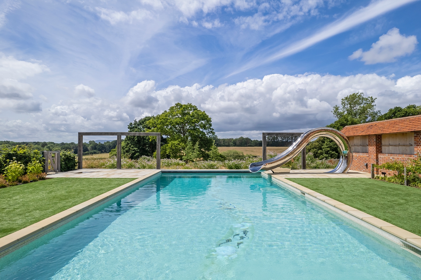 Panoramic image of outdoor swimming pool, with waha slide to the right, surronded by English countryside