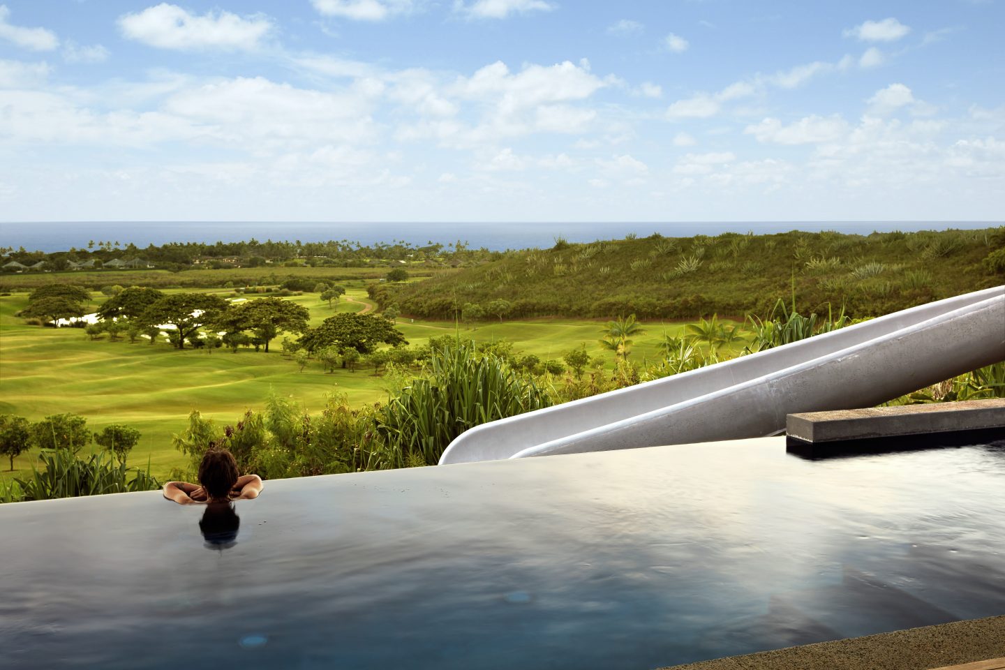 Concrete slide in front of infinity pool, with person on the pool looking at the view of Hawaii