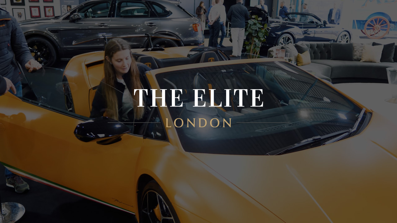 A high-end showroom featuring an orange luxury sports car in the foreground with "The Elite London" branding overlaid. In the background, other luxury vehicles, designer furniture, and event attendees are visible within an airplane hangar.