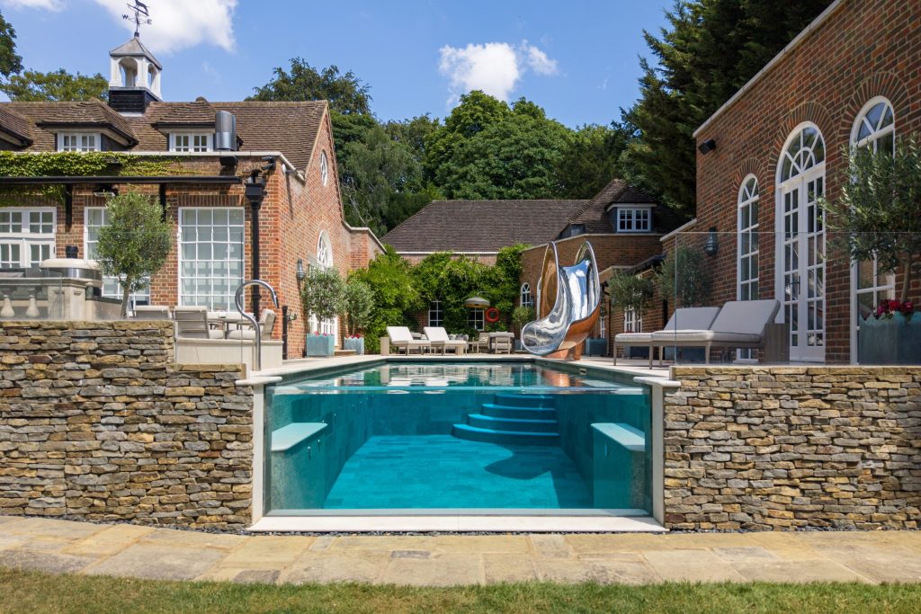 Compact stainless steel pool slide with a polished copper lustre finish beside a glass-fronted luxury garden pool in Surrey, UK. Compact stainless steel pool slide with a polished copper lustre finish beside a glass-fronted luxury garden pool in Surrey, UK.