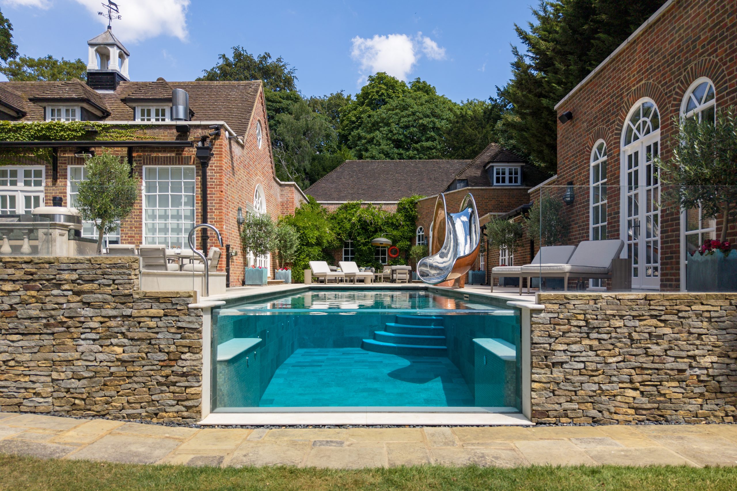 Compact stainless steel pool slide with a polished copper lustre finish beside a glass-fronted luxury garden pool in Surrey, UK.